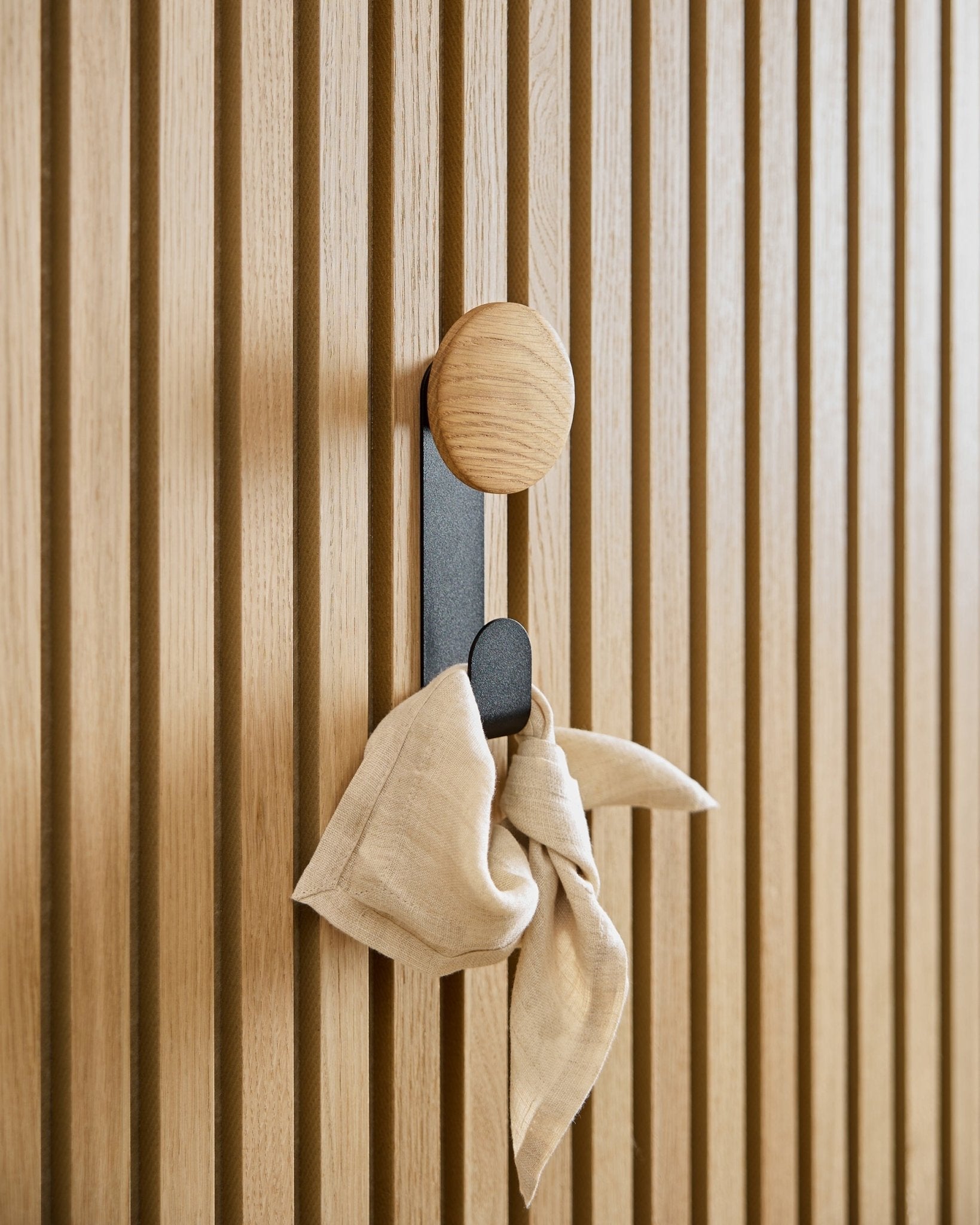 Un paño de beige cuelga del colgador Linn Vertical en Natural Oak de WoodUpp, montado sobre un panel acústico de listones vertical Akupanel.